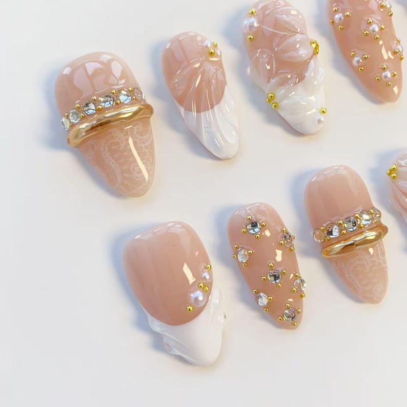 May include: A collection of artificial nails in various designs. Some nails are nude-colored with gold and rhinestone accents, while others feature white tips and pearl or floral embellishments. The nails showcase intricate lace patterns and a variety of decorative elements.