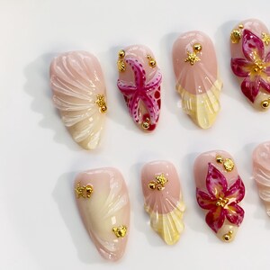 May include: A collection of artificial nails with a beach theme. The nails feature a variety of designs, including seashells, starfish, and floral accents. The color palette includes pink, white, yellow, and gold, creating a summery aesthetic.
