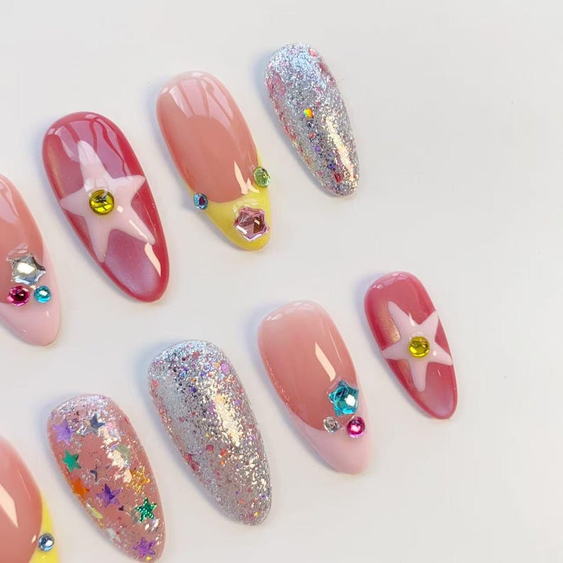 Pu&ograve; includere: Una collezione di unghie finte in vari design. Alcune unghie sono rosa, con un design a stella bianca e una gemma gialla. Altre sono ricoperte di glitter argentati o hanno una punta francese rosa e gialla con accenti di gemme.