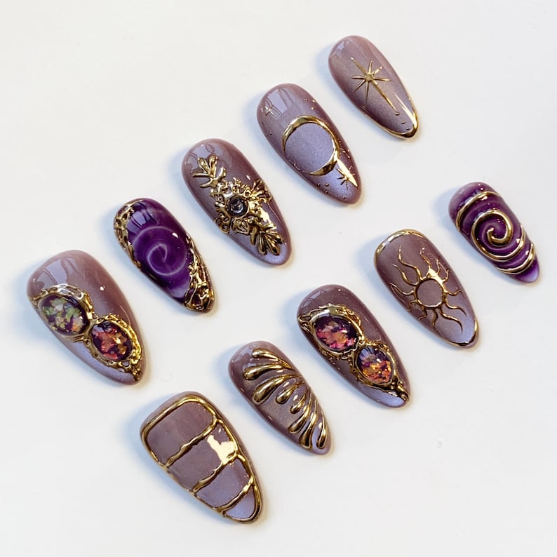 Pode incluir: Uma cole&ccedil;&atilde;o de unhas posti&ccedil;as em forma de am&ecirc;ndoa com v&aacute;rios designs. As unhas apresentam uma base roxa suave com detalhes dourados, incluindo motivos celestes como estrelas, luas e s&oacute;is, juntamente com redemoinhos ornamentados e enfeites de joias. A est&eacute;tica geral &eacute; elegante e art&iacute;stica.