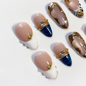 May include: Close-up of several artificial nails with various designs. Some nails are nude with gold accents, others feature a blue textured pattern, and some have a white textured design. The nails are oval-shaped and have a glossy finish.