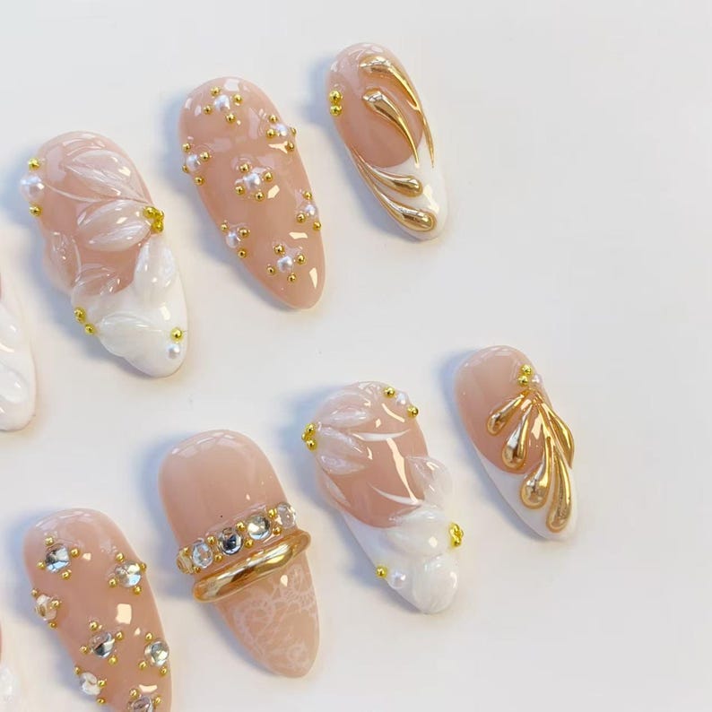 May include: A collection of almond-shaped artificial nails in various designs. Some nails feature a nude base with gold accents, pearls, and white floral designs. Others have gold rings with rhinestones or gold leaf patterns. The nails are arranged on a white surface.