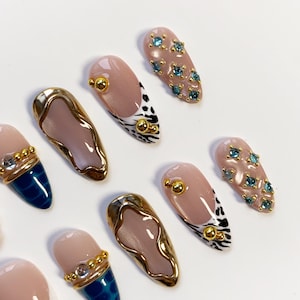 May include: A collection of artificial nails in various designs. Some nails are nude with gold accents, others feature blue and gold patterns, and some are adorned with blue gemstones. The nails are oval-shaped and have a glossy finish.