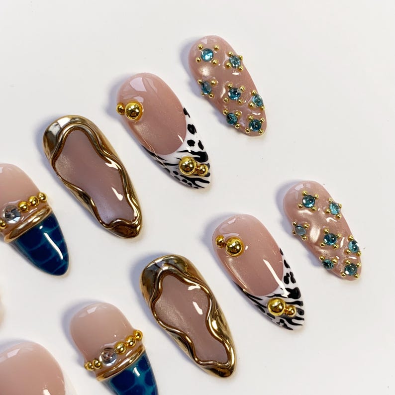May include: A collection of artificial nails in various designs. Some nails are nude with gold accents, others feature blue and gold patterns, and some are adorned with blue gemstones. The nails are oval-shaped and have a glossy finish.
