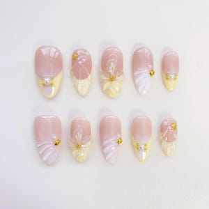 May include: A set of ten artificial nails in light pink and yellow. Each nail has a unique design, including floral accents, pearl embellishments, and gold-coloured studs. The nails have a glossy finish. The nails are designed for cosmetic use.