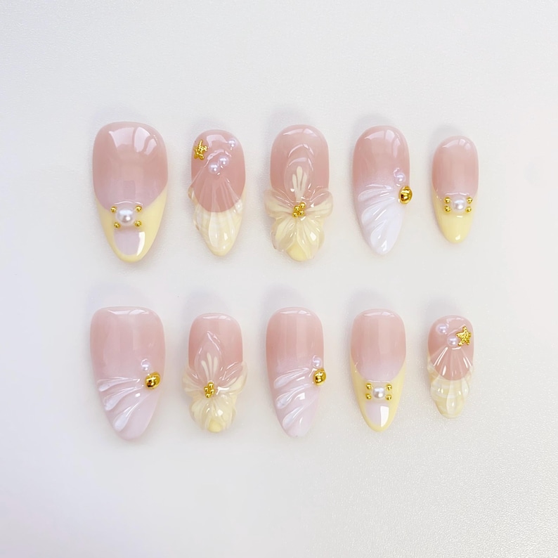 May include: A set of ten artificial nails in light pink and yellow. Each nail has a unique design, including floral accents, pearl embellishments, and gold-coloured studs. The nails have a glossy finish. The nails are designed for cosmetic use.