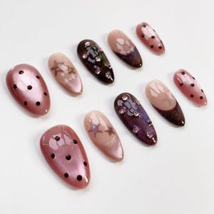 May include: A collection of artificial nails in various designs. Some nails are a shimmery pink with black polka dots. Others feature star designs, rhinestones, and ombre effects in shades of brown, pink, and purple. The nails are oval-shaped.