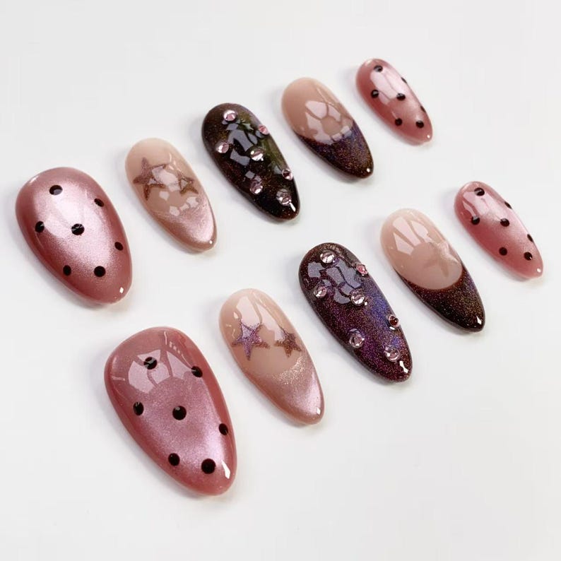 May include: A collection of artificial nails in various designs. Some nails are a shimmery pink with black polka dots. Others feature star designs, rhinestones, and ombre effects in shades of brown, pink, and purple. The nails are oval-shaped.