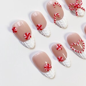 May include: A set of almond-shaped false nails with a nude base and white textured tips. The nails are decorated with red and white candy cane and peppermint designs, including a heart-shaped candy cane and a bow.