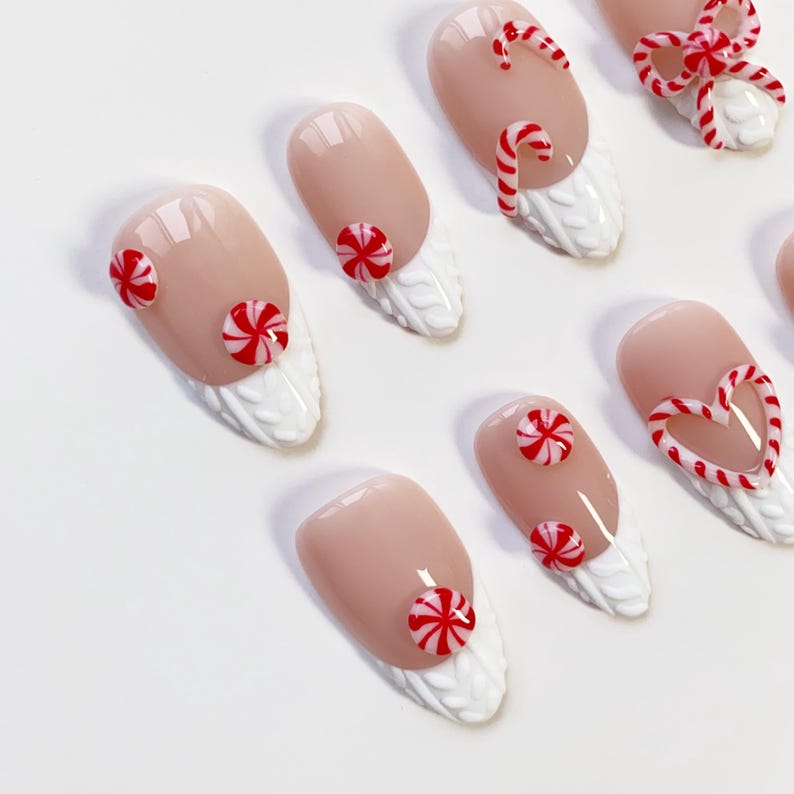 May include: A set of almond-shaped false nails with a nude base and white textured tips. The nails are decorated with red and white candy cane and peppermint designs, including a heart-shaped candy cane and a bow.