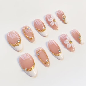 Puede incluir: Un juego de diez u&ntilde;as postizas con base color nude y puntas blancas. Los dise&ntilde;os incluyen detalles de filigrana dorada, peque&ntilde;os diamantes de imitaci&oacute;n transparentes y adornos florales blancos en relieve en dos de las u&ntilde;as almendradas.