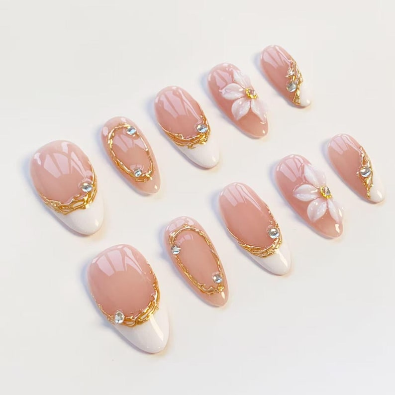 Puede incluir: Un juego de diez u&ntilde;as postizas con base color nude y puntas blancas. Los dise&ntilde;os incluyen detalles de filigrana dorada, peque&ntilde;os diamantes de imitaci&oacute;n transparentes y adornos florales blancos en relieve en dos de las u&ntilde;as almendradas.