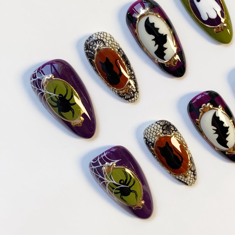 May include: A collection of Halloween-themed false nails. The nails feature designs of spiders, bats, and black cats in ornate frames, with a color palette of purple, black, white, and gold. Some nails have a spiderweb design.