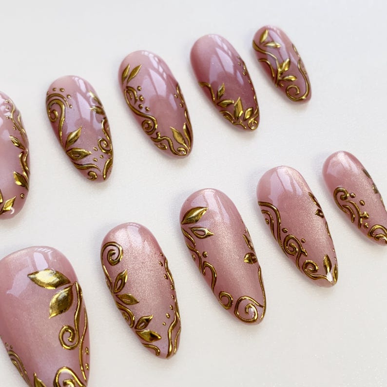 May include: A set of almond-shaped false nails in a soft pink hue, adorned with intricate gold leaf and swirl designs. The nails are arranged on a white surface, showcasing the detailed artistry and elegant aesthetic.