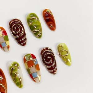 K&ouml;nnte beinhalten: Eine Sammlung k&uuml;nstlicher N&auml;gel in verschiedenen Designs. Einige N&auml;gel zeigen eine Spirale aus Golddraht &uuml;ber einem tiefroten Untergrund. Andere haben ein Mosaikmuster aus bunten Quadraten. Mehrere N&auml;gel haben goldene Verzierungen und ein gl&auml;nzendes Finish.