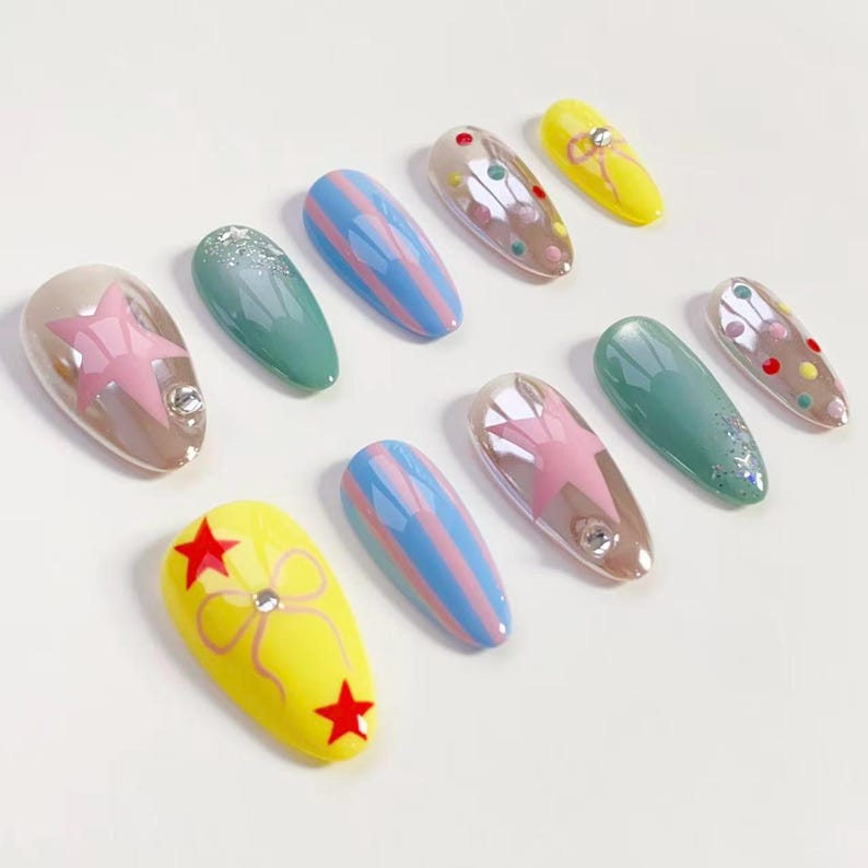 K&ouml;nnte beinhalten: Eine Sammlung k&uuml;nstlicher Fingern&auml;gel in verschiedenen Designs. Einige sind gelb mit roten Sternen, andere schimmern mit rosa Sternen, und einige sind t&uuml;rkis oder blau mit Streifen. Jeder Nagel hat ein gl&auml;nzendes Finish.