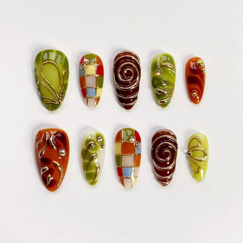K&ouml;nnte beinhalten: Ein Set aus zehn k&uuml;nstlichen N&auml;geln mit verschiedenen Designs. Die N&auml;gel weisen Farben wie Gr&uuml;n, Rot, Orange und Gelb auf, mit Golddraht und Edelsteinakzenten. Einige N&auml;gel haben Wirbelmuster, andere Gitter- oder Blumendesigns.