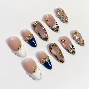 May include: A collection of ten artificial nails with various designs. Some nails are nude with gold accents, others feature blue and gold details, and some have a white and gold design. Several nails have blue gemstones.