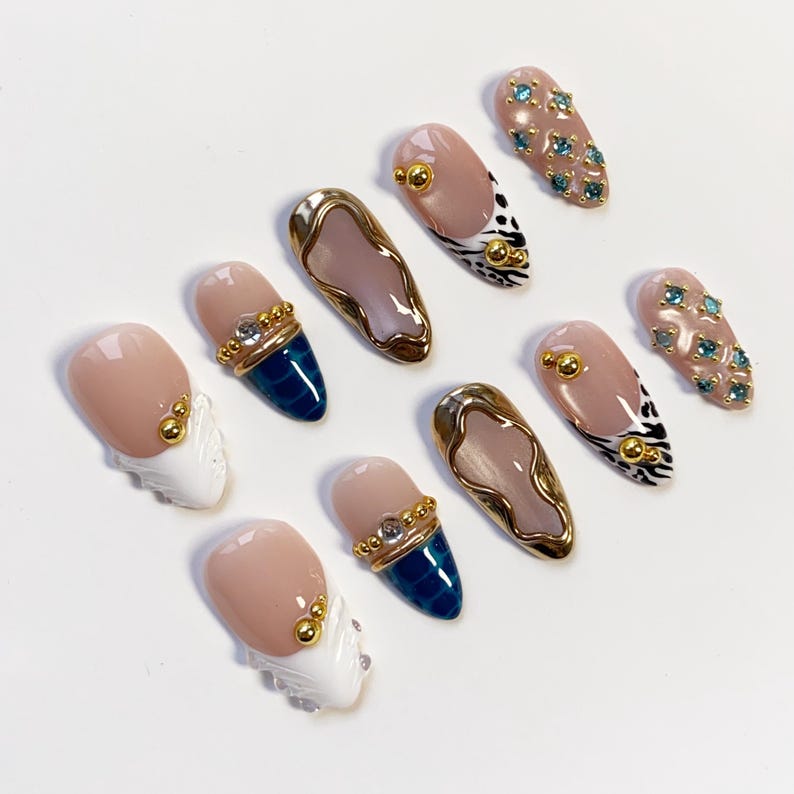 May include: A collection of ten artificial nails with various designs. Some nails are nude with gold accents, others feature blue and gold details, and some have a white and gold design. Several nails have blue gemstones.