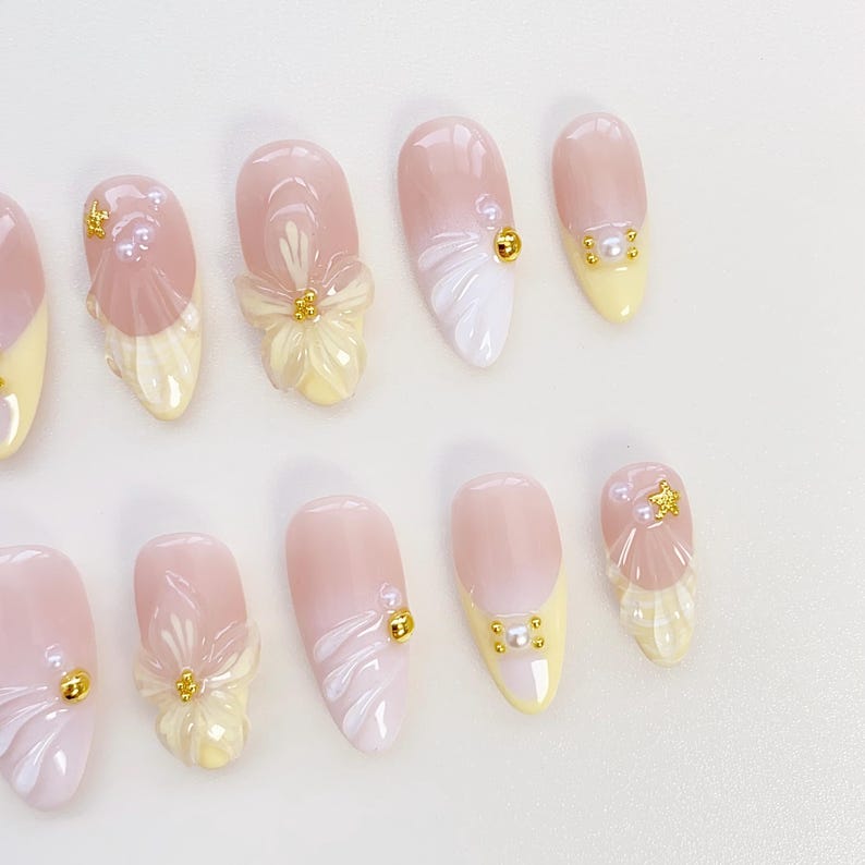 May include: A collection of almond-shaped false nails. The nails have a light pink base with a yellow tip. Some nails are decorated with 3D floral and pearl details, while others feature gold-coloured embellishments.
