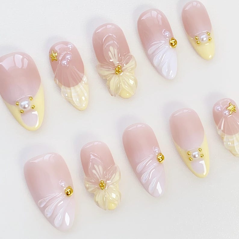 May include: A collection of ten artificial nails in a light pink and yellow palette. Each nail showcases a distinct design, incorporating floral details, pearl adornments, and gold-toned studs. The nails have a glossy finish.