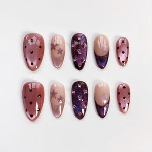 May include: A set of ten artificial nails in various designs. Some nails are a mauve color with black polka dots, others feature a star design, and some are a deep purple with rhinestones. The nails have an oval shape.