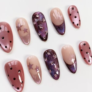 May include: A collection of ten artificial nails in various designs. Some nails are a shimmery pink with black polka dots, others feature a gradient of pink and purple with star accents, and some have rhinestones.