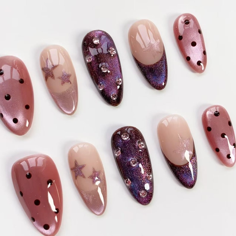 May include: A collection of ten artificial nails in various designs. Some nails are a shimmery pink with black polka dots, others feature a gradient of pink and purple with star accents, and some have rhinestones.