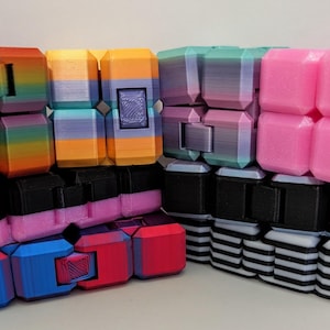 May include: A collection of eight multi-colored infinity cubes arranged in a group. The cubes feature various patterns including solid pink, rainbow gradients, horizontal stripes, and blue and red color blocks, all constructed from plastic segments.