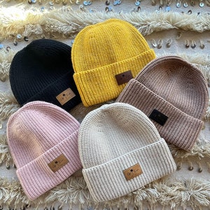 May include: Four knit beanies in different colors: black, yellow, pink, and beige. Each beanie has a small leather patch with the text "Off-Kai" and a palm tree design.