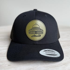 May include: Black baseball cap with a gold emblem featuring a building and Arabic text. The cap has a curved brim and a snapback closure. The emblem is circular and prominently displayed on the front of the cap.