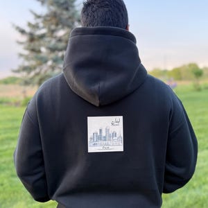 May include: Black hoodie with a square graphic on the back featuring a skyline and text in a foreign language. The hoodie has a large hood and is made of a thick material. The person is standing in a grassy field.