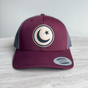 May include: A burgundy trucker hat with a mesh back and a curved brim. The front features a circular patch with a crescent moon and star design in gold and black. The hat has a snapback closure.