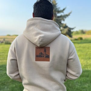May include: A beige hoodie with a hood, featuring a brown square patch on the back with the word "SYRIA" and a detailed illustration of a building. The hoodie is worn outdoors on a sunny day.