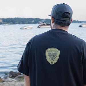 May include: A person wearing a black t-shirt and a dark gray baseball cap, standing near a lake with boats in the background. The t-shirt features a gold shield design with the word "PALESTINE" and a geometric pattern.