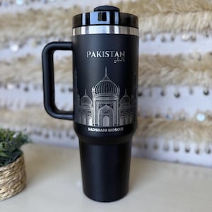 May include: Black travel mug with a handle, featuring the word "PAKISTAN" and the Urdu script, along with an etched image of the Badshahi Mosque. The mug has a silver rim and a black lid.