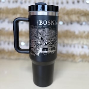 May include: Black travel mug with a handle and a black lid. The mug features the word "BOSNIA" and a detailed etched illustration of a cityscape. The word "SARAJEVO" is etched at the bottom of the mug.