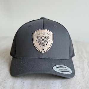 May include: A gray trucker hat with a mesh back and a curved brim. The hat features a tan leather patch with the word "PALESTINE" above a keffiyeh design. A "Snapback" tag is visible.