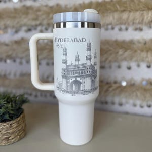 May include: White travel mug with a handle, featuring a gray illustration of the Charminar monument in Hyderabad. The mug has a silver rim and a white lid. The word "HYDERABAD" is printed above the illustration.