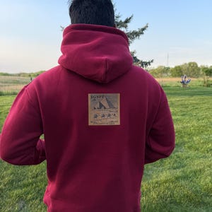 May include: A burgundy hoodie with a hood, featuring a small rectangular graphic on the back. The graphic depicts an illustration of the pyramids of Giza with the word "EGYPT" above and "PYRAMIDS OF GIZA" below.