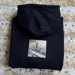 May include: Black hoodie with a hood. A rectangular patch on the front features a black and white illustration of a cityscape with the word "IRAQ" and Arabic text. The hoodie is made of a soft material.