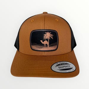 May include: A brown trucker hat with a black mesh back and a curved brim. The front features a rectangular patch with a camel and palm tree design. The hat has a snapback closure and a silver tag that reads "Yupoong Since 1974."
