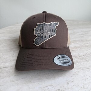 May include: A brown trucker hat with a tan mesh back and a curved brim. The front features a patch with a detailed illustration of a city within the outline of a country. The hat has a snapback closure and a "Snapback" tag.