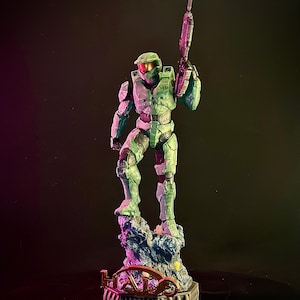 May include: An action figure of Master Chief from the Halo video game series. The figure is green and purple, holding a gun and standing on a rocky base with the word "HALO" written on it.