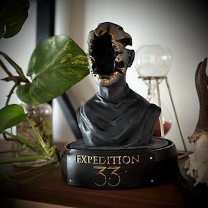 Może przedstawiać: Czarna rzeźba popiersia z wydrążoną głową i złotymi akcentami. Na podstawie napis "EXPEDITION 33" złotymi literami. Rzeźba stoi na drewnianej powierzchni, z rośliną i innymi elementami dekoracyjnymi w tle.