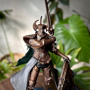 May include: A bronze-colored figurine of a knight in full armor, holding a sword and shield. The knight's helmet has curved horns, and the armor is intricately detailed. A green cape hangs from the knight's shoulders. The shield has a wolf design.