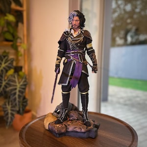 Puede incluir: Figura detallada de un hombre con un atuendo negro y morado, sosteniendo una espada. La figura tiene cabello negro con una raya blanca y está de pie sobre una base rocosa. El atuendo incluye detalles dorados y una faja morada.