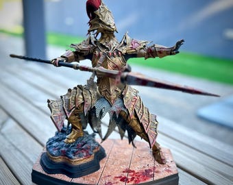 May include: A detailed figurine of a warrior in ornate gold armor, holding a spear and a blood-stained sword. The warrior is posed in a dynamic stance on a base with a textured surface, featuring red accents. The helmet has a red plume.