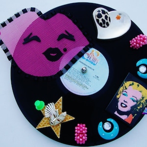 May include: A black vinyl record with various colorful embellishments, including a pink and black lips design, a silver heart with a black and white animal print, a gold star with a white bird design, a green push pin, and a small picture of Marilyn Monroe. The record label reads "20th Century" and "Remember Marilyn".