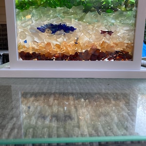 May include: A rectangular framed art piece filled with layers of sea glass. The top layer is green, followed by light green, white, blue, orange, and brown. The frame is white.